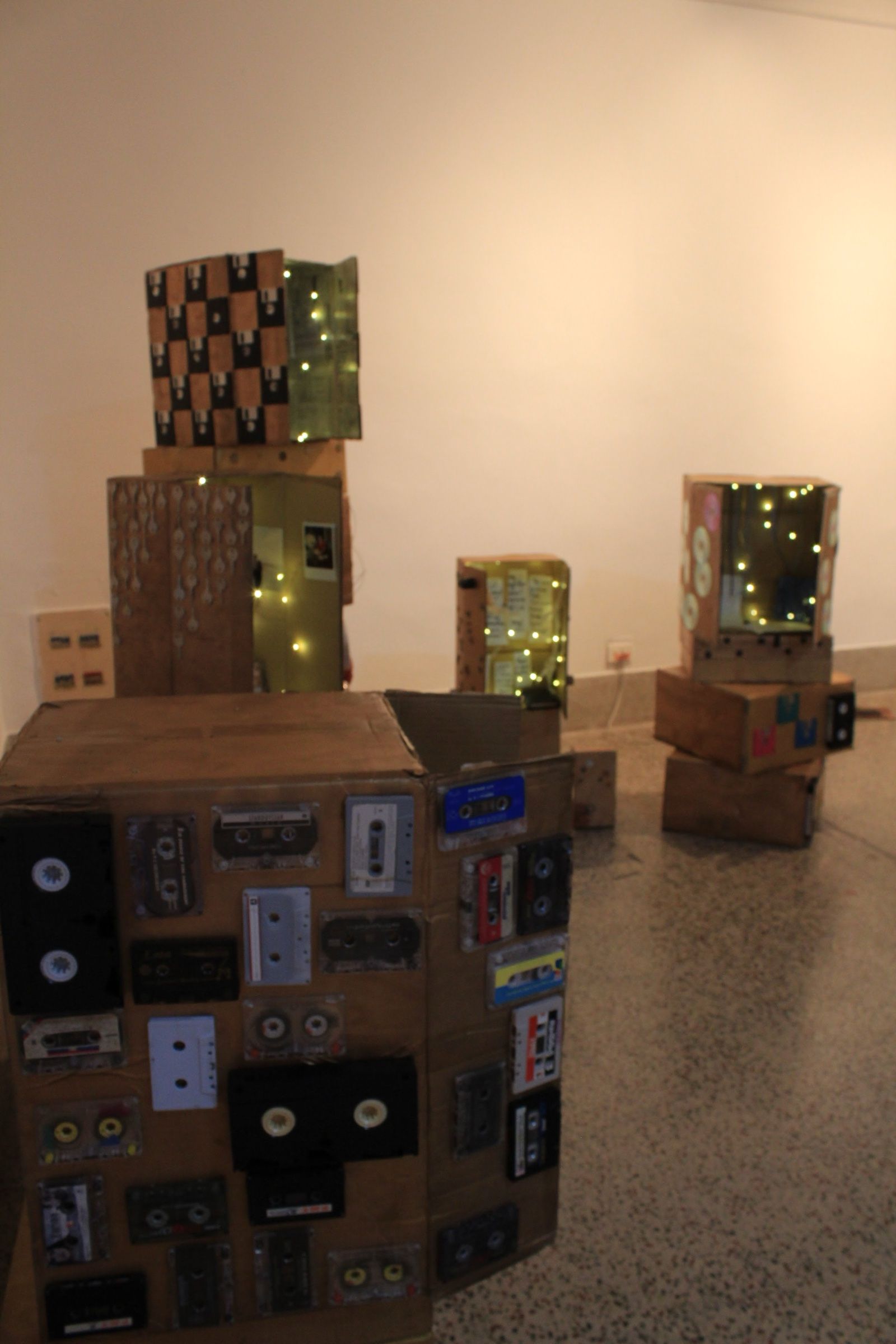 A cardboard shrine covered with cassette tapes, with illuminated structures behind it.
